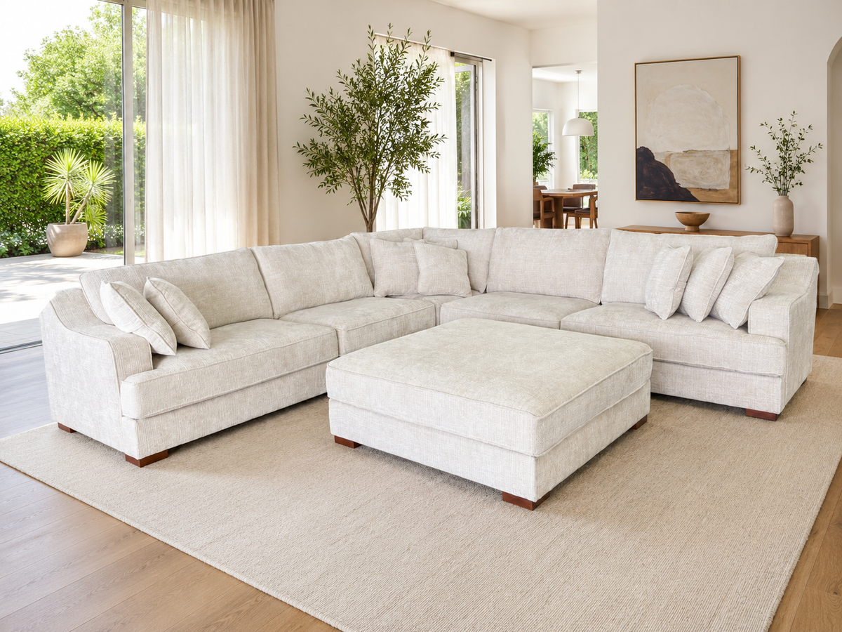 A large U-shaped sectional sofa in a light beige textured fabric, paired with a matching square ottoman, set on a large beige area rug in a bright, modern living room.