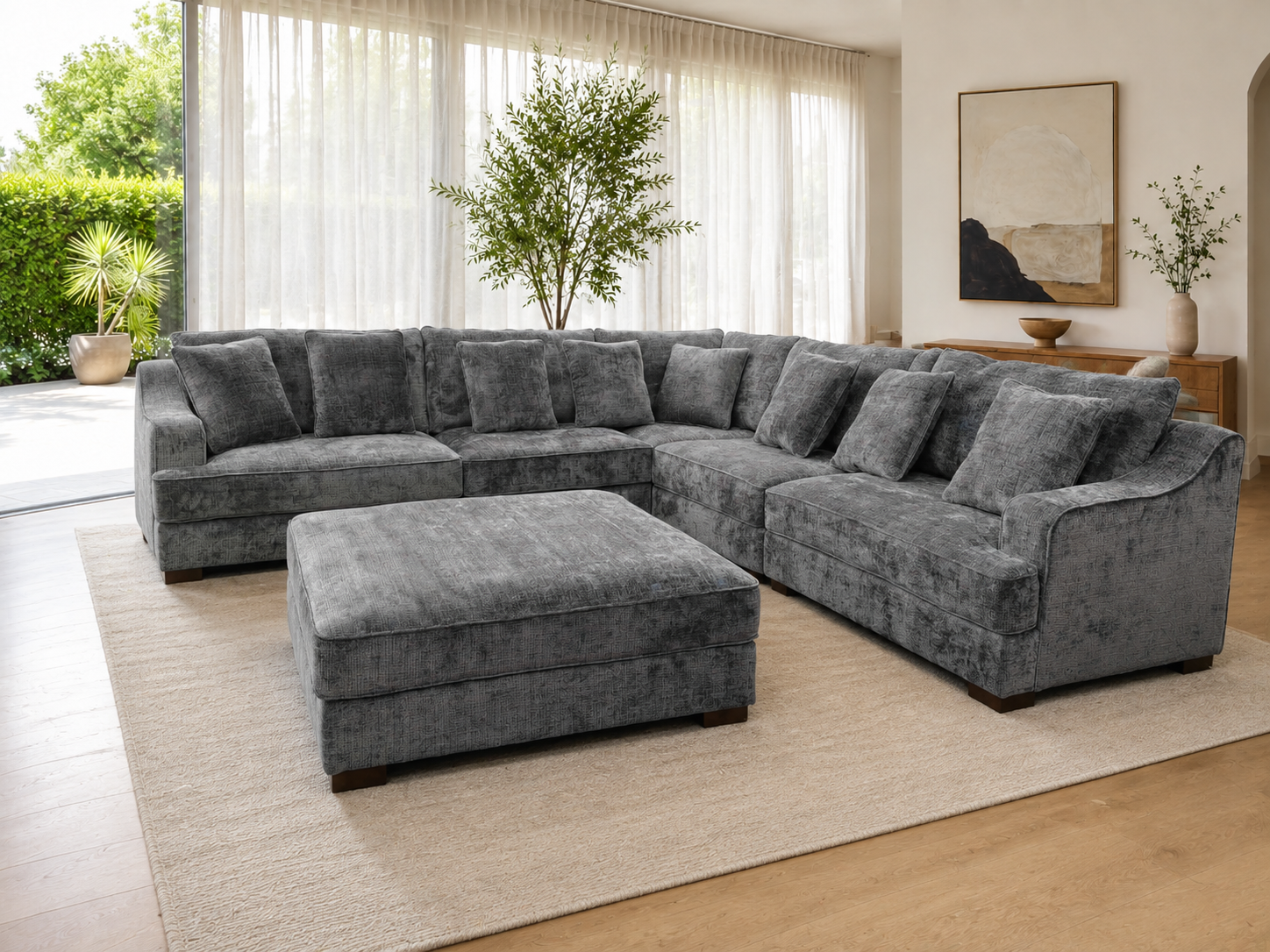 A large U-shaped sectional sofa in textured charcoal gray velvet, paired with a matching square ottoman, set on a beige area rug in a bright, modern living room.